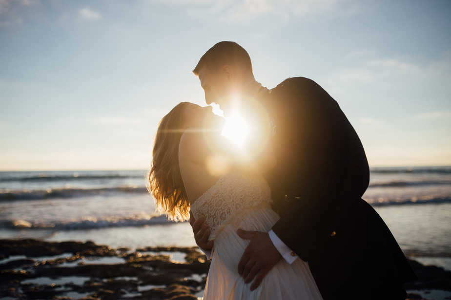 Bride and groom stand on the beach during sunset with sun flare